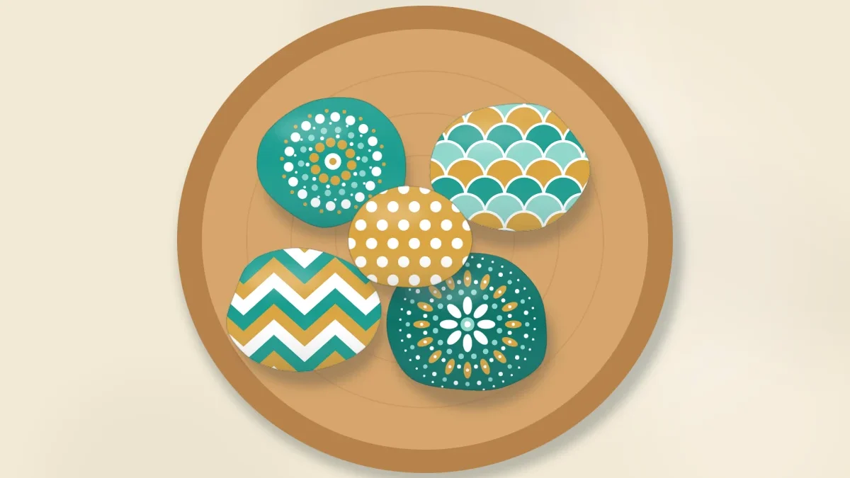 A round wooden bowl seen from above filled with decorative painted stones in a matching teal, white and gold palette