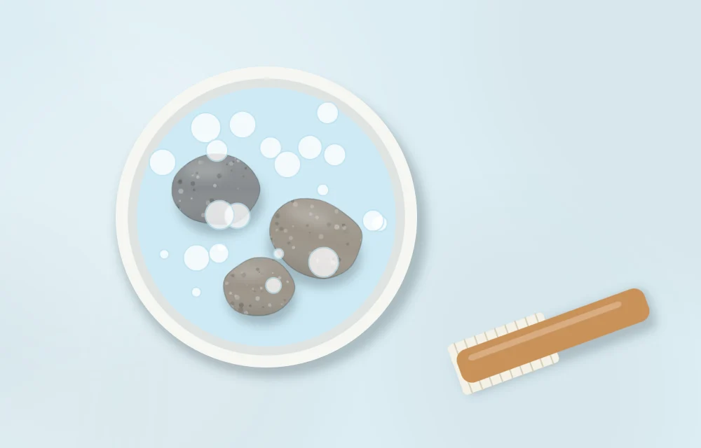 Round bowl of soapy water seen from above with three stones in it and a small scrub brush lying next to the bowl